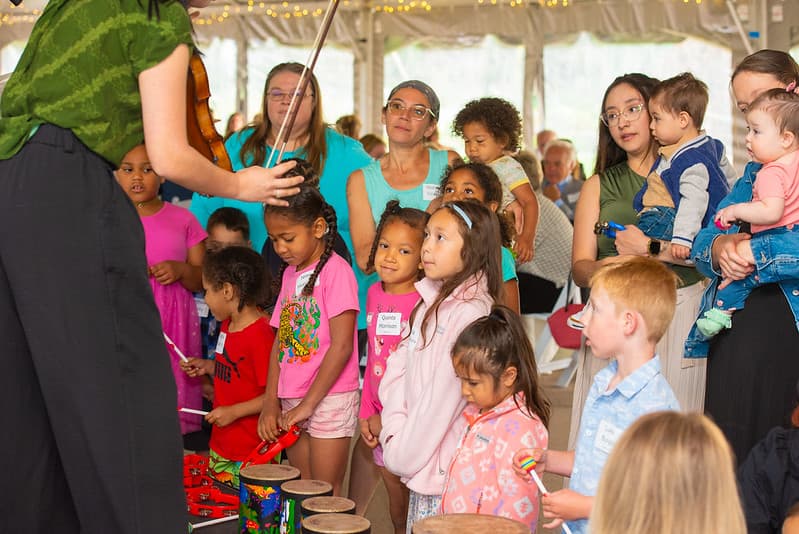 A violinist plays while young children drum and shake percussion instruments as families watch under a tent at Stone Soup.