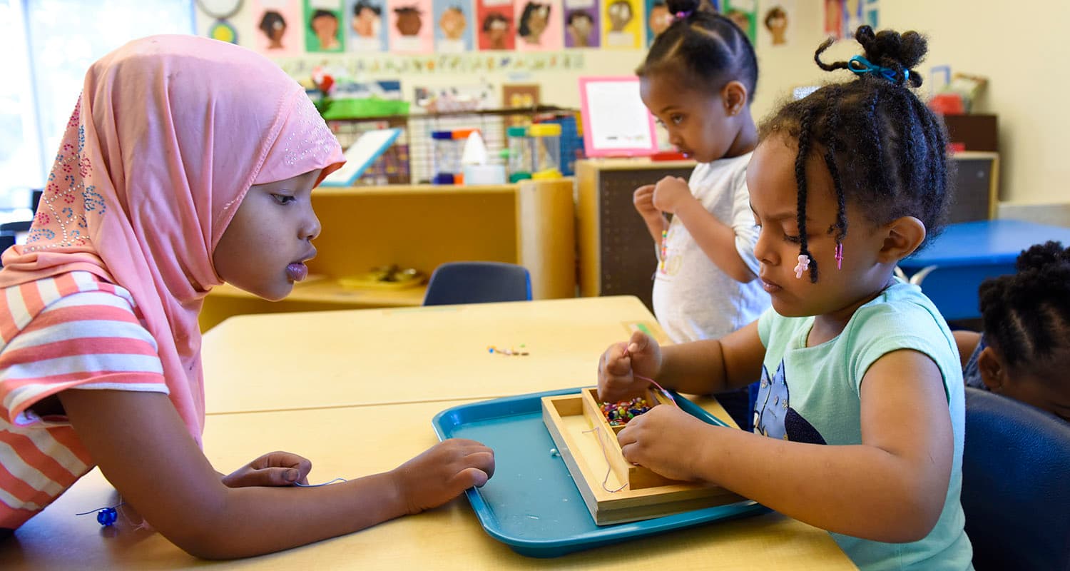 Two young girls, one in a pink hijab, threading beads onto laces from a Montessori tray.